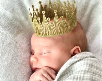 May include: A gold glittery crown with a pointed, open-work design featuring circular tips. The crown is constructed from a stiffened, metallic-threaded fabric that forms a series of triangular peaks, creating a structured, circular headpiece.