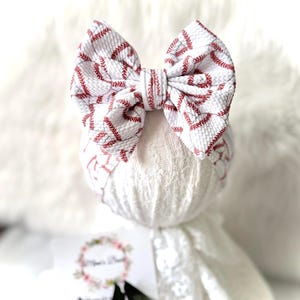 May include: A white fabric hair bow featuring a repeating red baseball stitch pattern. The bow has a textured, seersucker-like surface and is centered on a matching white fabric headband. The design includes a central knot detail made of the same fabric.