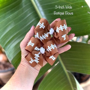 May include: Brown football-themed boutique bows, each 3 inches wide, with white stitching details. The bows are held in a hand against a backdrop of green leaves. The text "3-Inch Wide Botique Bows" is visible.