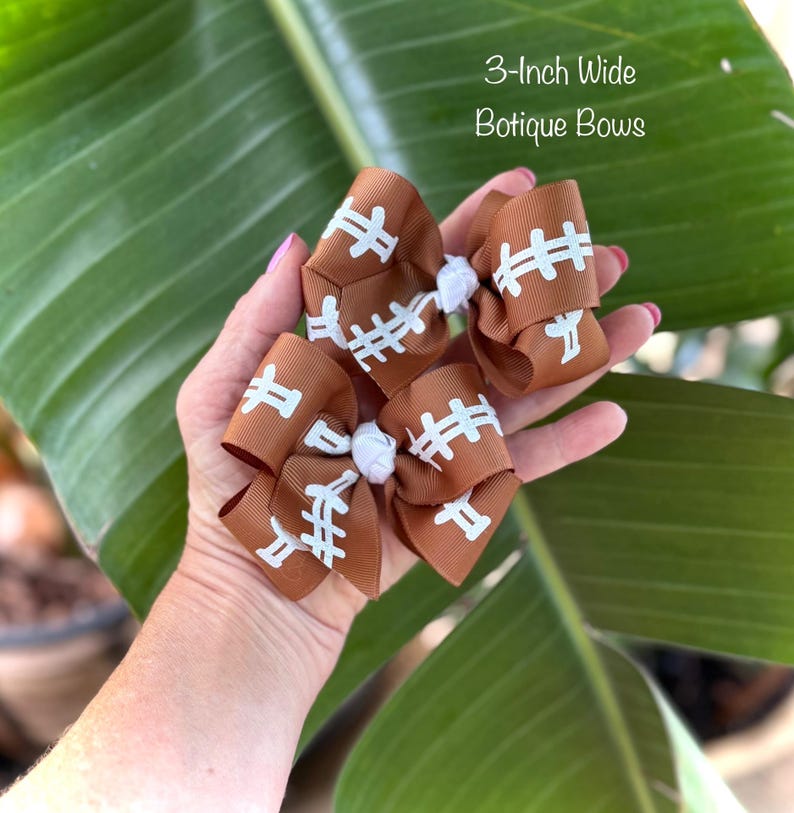 May include: Brown football-themed boutique bows, each 3 inches wide, with white stitching details. The bows are held in a hand against a backdrop of green leaves. The text "3-Inch Wide Botique Bows" is visible.