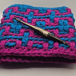 Könnte beinhalten: Ein handgefertigtes Häkelprojekt in leuchtendem Blau und Magenta. Das strukturierte Stück hat ein gewebtes Muster, mit einem dunkelbraunen Häkelhaken, der darauf liegt. Der Haken hat einen hellen Griff und ein spitzes Ende, was auf detaillierte Arbeit hindeutet.