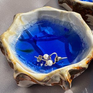 Op de afbeelding: Een kleine, onregelmatige komvormige schaal met een glanzende, crèmekleurige en bruin gemarmerde rand. De binnenkant is gevuld met een levendige, doorschijnende blauwe hars die water nabootst, met daarin één goudkleurige oorbel met twee witte parels.