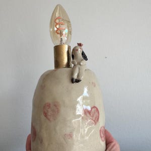 May include: A ceramic table lamp with a cream-coloured, textured base featuring hand-painted pink hearts. A small, sculpted figure with dark hair sits on the rim. A gold-toned socket holds a clear, spiral-filament light bulb at the top.