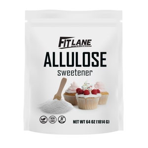 May include: A white bag of Fit Lane Nutrition Allulose sweetener, a sugar substitute. The bag is labeled with the product name, "Allulose Sweetener", and the net weight, "64 oz (1814 g)". The bag also features a vegan logo, a non-GMO logo, and a gluten-free logo. The bag is shown with a wooden spoon scooping out the white powder and three cupcakes with pink and white frosting.