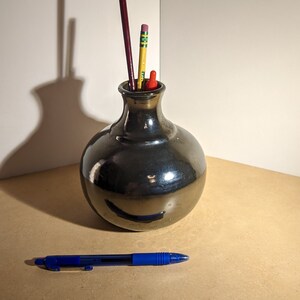 May include: A black ceramic vase with a round body and a narrow neck holds a paintbrush and three pencils. A blue pen is lying on a light brown surface.