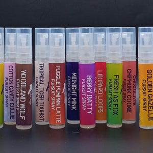 May include: A collection of twelve spray bottles with clear caps and colorful labels. Each label features a unique name, such as "Sour Spinosaur" and "Lavender Lynx", along with the text "Fursuit Spray". The bottles are arranged in a row.