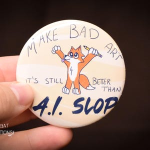May include: A circular badge featuring a cartoon fox with a pencil, and the text "MAKE BAD ART" and "IT'S STILL BETTER THAN A.I. SLOP". The fox is orange and white. The badge is held in a hand.