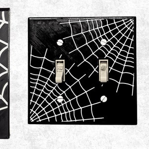 Könnte beinhalten: Zwei schwarze Lichtschalterabdeckungen mit einem weißen Spinnennetz-Design. Die Schalterabdeckungen sind für Standard-Lichtschalter.