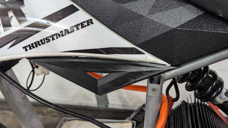 Puede incluir: Primer plano de un simulador de carreras blanco y negro con la palabra "THRUSTMASTER" impresa en el lateral. El simulador tiene un volante negro y gris y un asiento negro y gris.