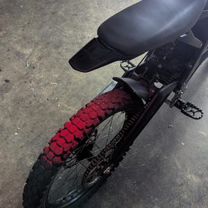 May include: Black electric dirt bike with a red-painted rear tire. The bike has a black seat, fenders, and frame. The tire has a knobby tread pattern. The bike is designed for off-road use.