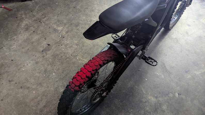 May include: Black electric dirt bike with a red-painted rear tire. The bike has a black seat, fenders, and frame. The tire has a knobby tread pattern. The bike is designed for off-road use.