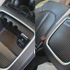 May include: A black car console with a silver trim. The console has a storage compartment, cup holders, and a sliding cover with a ridged design. The interior is black leather.