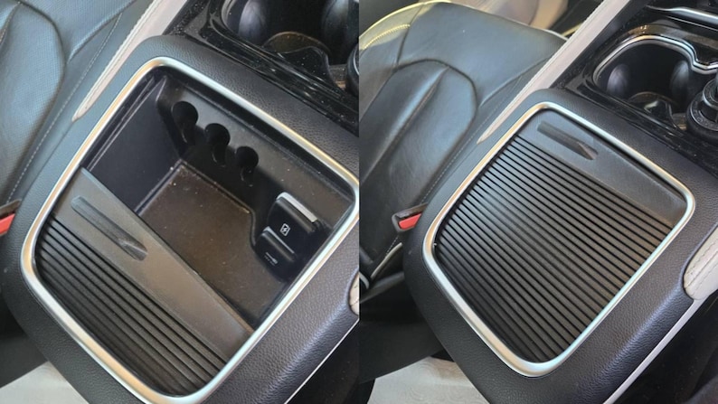 May include: A black car console with a silver trim. The console has a storage compartment, cup holders, and a sliding cover with a ridged design. The interior is black leather.
