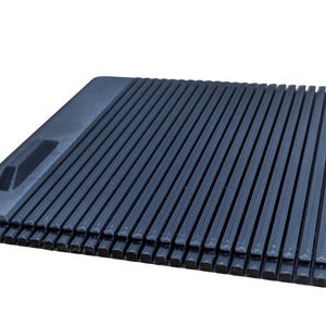 May include: A dark gray, rectangular object with a ridged surface. The object has a raised section on one end with a rectangular cutout. The ridges run parallel to each other, creating a textured appearance. The object is likely made of plastic.
