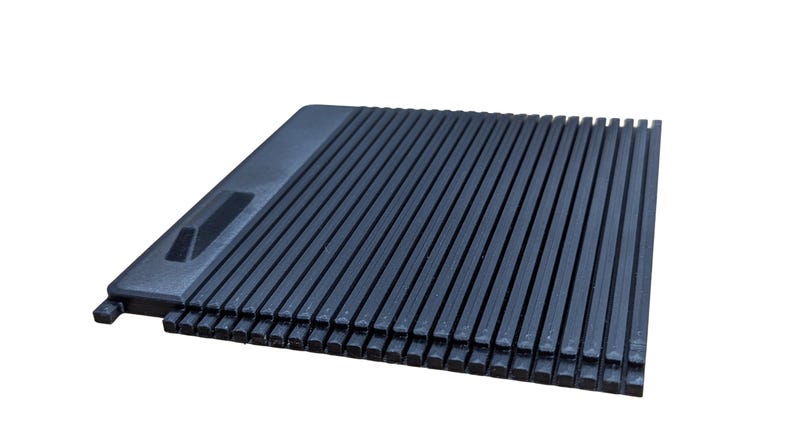 May include: A dark gray, rectangular object with a ridged surface. The object has a raised section on one end with a rectangular cutout. The ridges run parallel to each other, creating a textured appearance. The object is likely made of plastic.