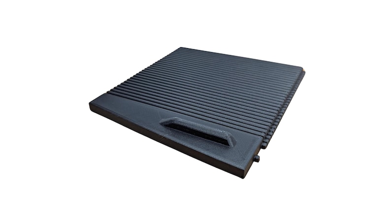 May include: A black, rectangular grill grate with a handle on one side. The grate features parallel ridges across its surface. The handle is a recessed, rectangular shape. The item is made of a dark, matte material.