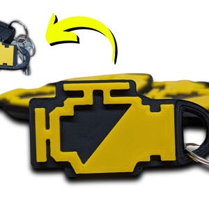 Może przedstawiać: Żółto-czarny brelok do kluczy w kształcie kontrolki silnika. Brelok ma kształt symbolu kontrolki silnika. Brelok jest przymocowany do srebrnego kółka na klucze.