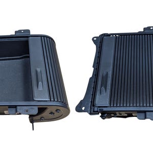 May include: Two black plastic car console components are displayed against a white background. The components have a rectangular shape with a ribbed design on the front and a storage compartment. The components are likely for a vehicle's interior.