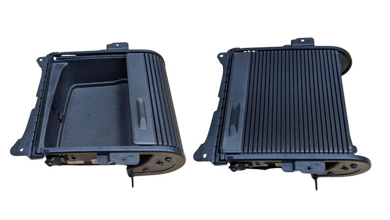May include: Two black plastic car console components are displayed against a white background. The components have a rectangular shape with a ribbed design on the front and a storage compartment. The components are likely for a vehicle's interior.