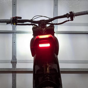 May include: Rear view of a black motorcycle with a bright red taillight. The handlebars are visible, along with the grips and controls. The motorcycle is parked in front of a white garage door.