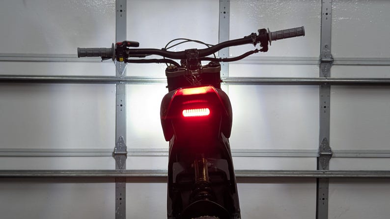 May include: Rear view of a black motorcycle with a bright red taillight. The handlebars are visible, along with the grips and controls. The motorcycle is parked in front of a white garage door.