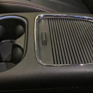 May include: Close-up of a black leather car interior with a chrome-trimmed cup holder and a black, ribbed, rectangular air vent.