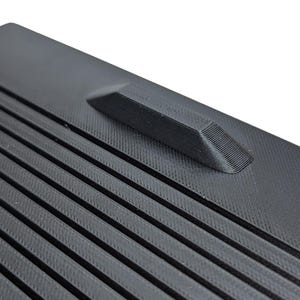 May include: Close-up of a black plastic surface with a textured, ribbed pattern. A small, raised, rectangular bump is located on the surface.