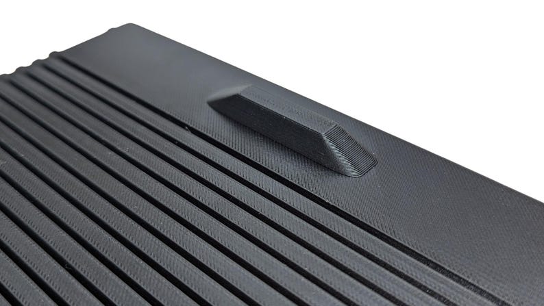 May include: Close-up of a black plastic surface with a textured, ribbed pattern. A small, raised, rectangular bump is located on the surface.