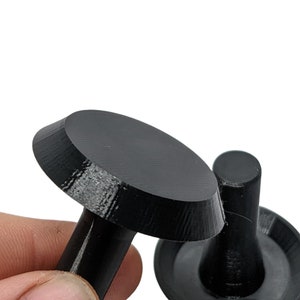 Op de afbeelding: Twee zwarte, 3D-geprinte objecten van plastic. Een object is een ronde schijf met een korte, cilindrische steel. Het andere object is een korte, cilindrische steel met een ronde schijfbasis.