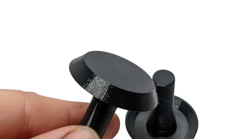 Op de afbeelding: Twee zwarte, 3D-geprinte objecten van plastic. Een object is een ronde schijf met een korte, cilindrische steel. Het andere object is een korte, cilindrische steel met een ronde schijfbasis.