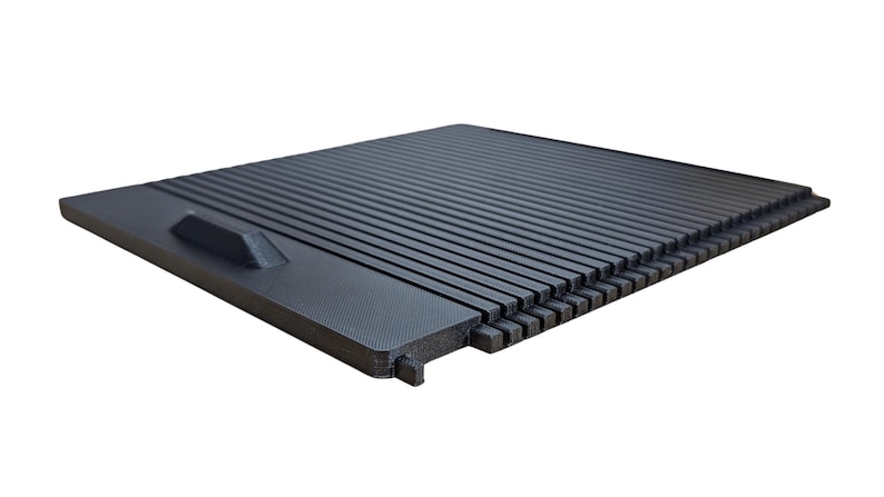 May include: A black plastic car part with a textured surface and a small handle. The part has a series of parallel lines and a grid pattern.