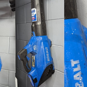 K&ouml;nnte beinhalten: Zwei blaue und schwarze kabellose Laubbl&auml;ser, die an einer Wand h&auml;ngen. Die Gebl&auml;se sind mit "40V MAX" und "KOALT" beschriftet und haben einen Aufkleber mit der Aufschrift "480 CFM 110 MPH".
