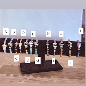 May include: A collection of beaded bracelets with seashell accents. The bracelets feature various colors of beads, including blue, brown, and white, and are displayed on a black stand. Each bracelet has a unique seashell design.