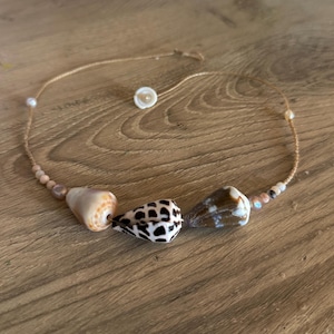 May include: A necklace featuring natural seashells, pearls, and small beads strung on a delicate cord. The shells vary in color, including white, brown, and black, with a unique spotted pattern on one shell. The necklace is displayed on a wooden surface.