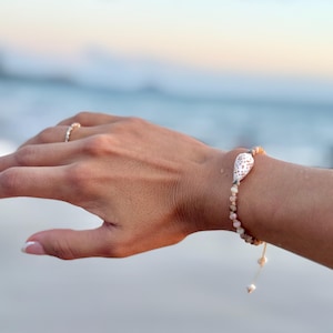Puede incluir: Una pulsera de conchas con pequeñas cuentas claras y una concha blanca más grande en el centro. La pulsera se lleva en la muñeca, con un anillo en un dedo. El fondo es una vista borrosa del océano.