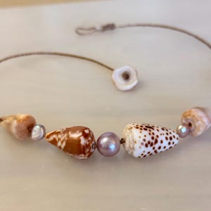 May include: A delicate bracelet featuring a thin brown cord strung with a central smooth lavender pearl, flanked by two distinct cone shells—one solid brown and one white with brown spots—and two smaller tan shells with tiny grey pearls.