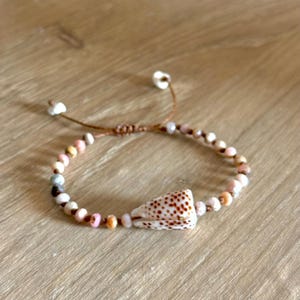 May include: A beaded bracelet featuring a natural seashell centerpiece. The bracelet is strung on a brown cord and includes small, colorful beads in shades of pink, white, and gray. The seashell has a speckled pattern.