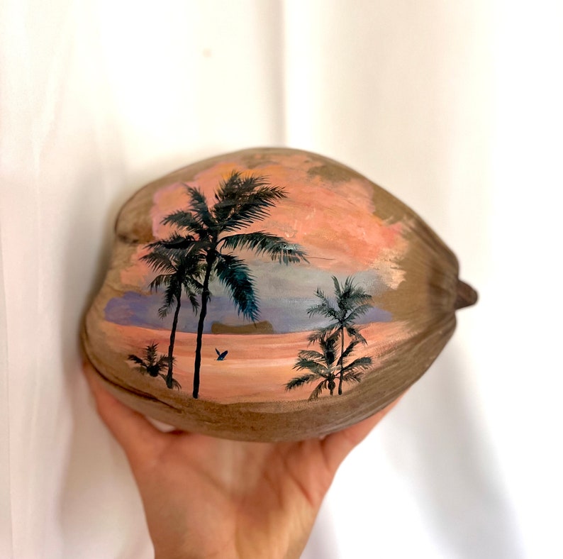 Puede incluir: Una c&aacute;scara de coco pintada a mano con una escena de puesta de sol tropical. La obra de arte representa palmeras contra un cielo rosa y naranja, con un toque de azul y un peque&ntilde;o p&aacute;jaro en vuelo. La c&aacute;scara de coco es marr&oacute;n.