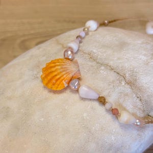May include: A necklace with an orange seashell pendant, alongside pink and white beads. The necklace is strung on a thin, natural-coloured cord, creating a beachy, coastal aesthetic.