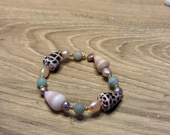 Könnte beinhalten: Ein handgefertigtes Armband mit einer Mischung aus natürlichen Muscheln, Perlen und Perlen. Das Armband enthält schwarz-weiß gefleckte Muscheln, hellrosa Perlen und kleine goldfarbene Perlen. Das Design beinhaltet hellblau-grüne Perlen und größere, hellrosa Muscheln.