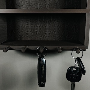 Peut inclure: Un porte-cl&eacute;s mural en bois brun fonc&eacute; dot&eacute; d'une &eacute;tag&egrave;re et de cinq crochets. Le panneau arri&egrave;re pr&eacute;sente un motif g&eacute;om&eacute;trique en forme d'&eacute;toile. Deux anneaux m&eacute;talliques avec des porte-cl&eacute;s noirs sont suspendus aux crochets inf&eacute;rieurs.