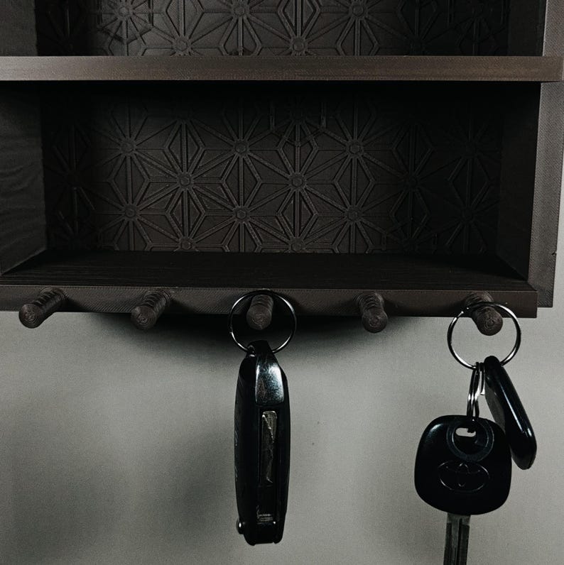 Peut inclure: Un porte-cl&eacute;s mural en bois brun fonc&eacute; dot&eacute; d'une &eacute;tag&egrave;re et de cinq crochets. Le panneau arri&egrave;re pr&eacute;sente un motif g&eacute;om&eacute;trique en forme d'&eacute;toile. Deux anneaux m&eacute;talliques avec des porte-cl&eacute;s noirs sont suspendus aux crochets inf&eacute;rieurs.