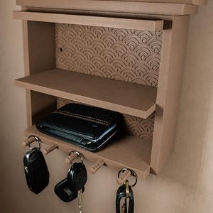 Peut inclure: Une &eacute;tag&egrave;re murale marron clair avec un sommet en forme de toit et un panneau arri&egrave;re textur&eacute; pr&eacute;sentant un motif de vagues r&eacute;p&eacute;titif. L'unit&eacute; comprend deux &eacute;tag&egrave;res horizontales et trois crochets en bois en bas, portant trois trousseaux de cl&eacute;s.