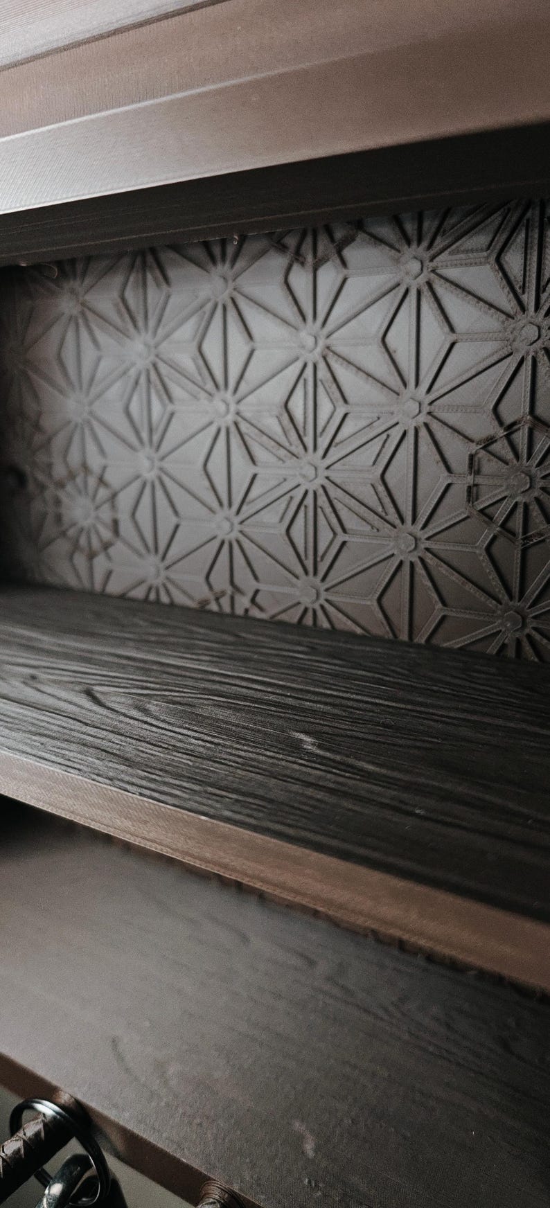 Peut inclure: Une &eacute;tag&egrave;re marron fonc&eacute; avec une texture de grain de bois, dot&eacute;e d'un panneau arri&egrave;re orn&eacute; d'un motif g&eacute;om&eacute;trique de triangles et de cercles r&eacute;p&eacute;t&eacute;s. L'&eacute;tag&egrave;re pr&eacute;sente une surface horizontale et une paroi arri&egrave;re verticale structur&eacute;e.