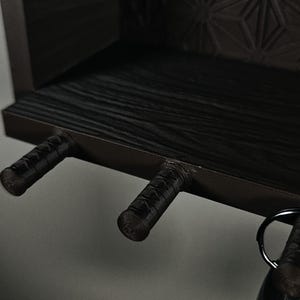 Peut inclure: Une &eacute;tag&egrave;re murale marron fonc&eacute; avec une surface en grain de bois textur&eacute; et un motif g&eacute;om&eacute;trique sur le panneau arri&egrave;re. L'&eacute;tag&egrave;re comprend trois crochets cylindriques saillants rappelant des poign&eacute;es d'&eacute;p&eacute;e, dont un porte un porte-cl&eacute;s.