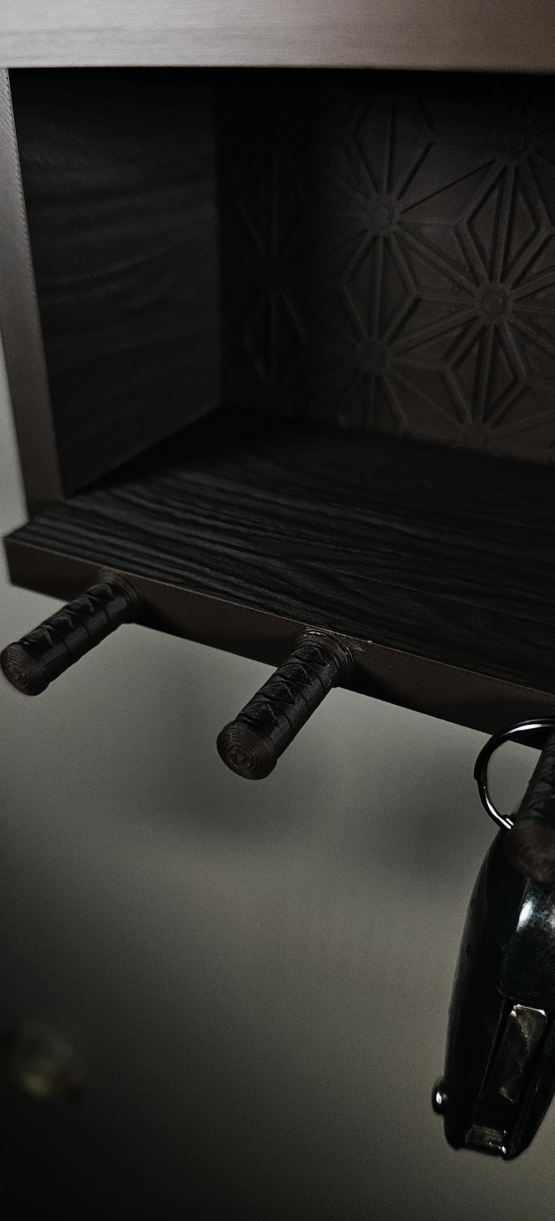 Peut inclure: Une &eacute;tag&egrave;re murale marron fonc&eacute; avec une surface en grain de bois textur&eacute; et un motif g&eacute;om&eacute;trique sur le panneau arri&egrave;re. L'&eacute;tag&egrave;re comprend trois crochets cylindriques saillants rappelant des poign&eacute;es d'&eacute;p&eacute;e, dont un porte un porte-cl&eacute;s.