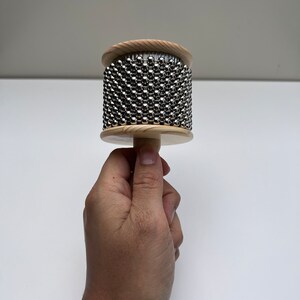 May include: A wooden percussion instrument with a cylindrical shape and a handle. The instrument is covered in small metal beads.
