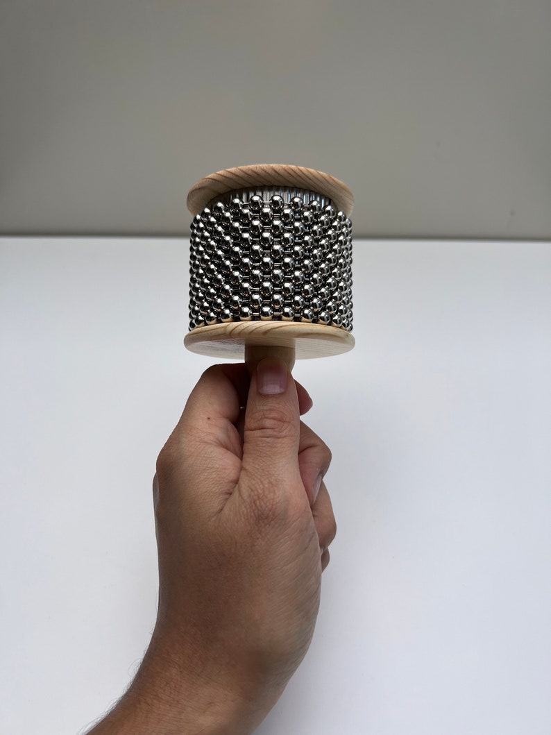 May include: A wooden percussion instrument with a cylindrical shape and a handle. The instrument is covered in small metal beads.