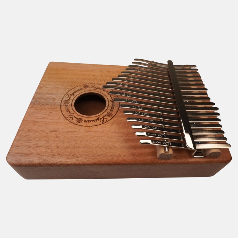 Puede incluir: Un kalimba de madera con un acabado de madera natural y l&aacute;minas de metal. El kalimba tiene un agujero de sonido circular y el texto "Signature Lyrewood Kalimba" est&aacute; grabado en el cuerpo.