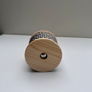 May include: A wooden percussion instrument with a circular shape and a band of metal beads around the center. The instrument is made of light wood and has a natural finish.